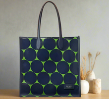Load image into Gallery viewer, A blue and green dotted Kate Spade Gotham Joy tote bag with leather trim and a visible logo on the front, placed on a surface with a plant in the background.