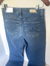 Load image into Gallery viewer, Back view of a pair of medium-wash blue denim jeans hanging on a hanger against a white wall, showing the back pockets with embroidered logo details.