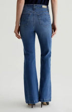 Load image into Gallery viewer, Rear view of a model wearing blue high-rise flare jeans with a dark top and black heels, set against a plain light gray studio background.