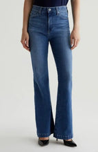 Load image into Gallery viewer, A model wearing a pair of medium-wash blue flare jeans with a high waist, paired with black pointed-toe heels against a light gray studio background.