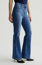 Load image into Gallery viewer, A model wearing a pair of medium-wash blue flare jeans with black pointed-toe heels, shown from the waist down against a light gray studio background.