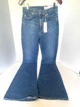 Load image into Gallery viewer, A pair of medium-wash blue denim flare jeans hanging on a wooden hanger against a white studio backdrop, with a white retail tag attached to the waistband.