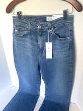 Load image into Gallery viewer, A pair of medium-wash blue denim jeans hanging from a wooden hanger with black clips, showing the front waistband, belt loops, button, pockets, and a white product tag.