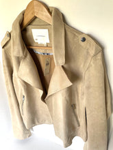 Load image into Gallery viewer, A light beige suede moto jacket hanging on a wooden hanger against a plain off-white wall. The jacket features an asymmetrical zipper, silver-toned snap buttons, and zippered pockets.