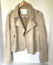 Load image into Gallery viewer, A light beige suede moto jacket hanging on a wooden hanger against a white wall. The jacket features a relaxed fit, shoulder epaulets, and zippered side pockets.