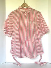 Load image into Gallery viewer, A short-sleeve collared shirt with vertical red and white stripes, hanging on a wooden hanger against a plain off-white wall. The shirt has a chest pocket and side ties at the hem.