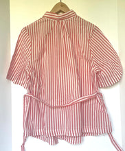 Load image into Gallery viewer, A pink and white vertically striped short-sleeve shirt with a tie waist belt, hanging on a wooden hanger against a plain off-white background.