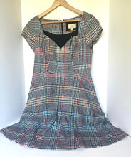 Load image into Gallery viewer, A vintage-style plaid dress with short sleeves and a black V-neckline, hanging on a wooden hanger against a white wall.