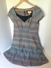 Load image into Gallery viewer, A vintage-style plaid dress with short cap sleeves and a gathered flared skirt, hanging on a wooden hanger against a white wall. The dress features a gray, black, and red plaid pattern with a solid black V-neckline.