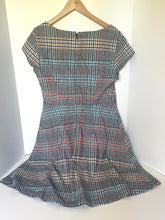 Load image into Gallery viewer, A short-sleeve plaid dress hanging on a wooden hanger against a white wall corner. The dress features a gray, black, white, and red plaid pattern.