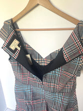 Load image into Gallery viewer, A sleeveless plaid top hanging on a wooden hanger against a plain light wall. The garment has a black V-neck trim and a multicolor plaid pattern with red, blue, and white lines on a dark base.