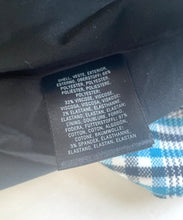 Load image into Gallery viewer, Close-up of a black fabric garment label with white text showing material composition and care instructions, placed on dark fabric with a blue-and-white plaid fabric visible at the lower left.
