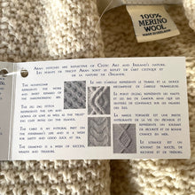 Load image into Gallery viewer, Close-up of a knitted fabric with a label and information sheet on merino wool and Aran stitches.