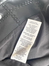 Load image into Gallery viewer, Close-up of a black leather jacket interior showing a white product care label with text details, alongside black leather trim and a silver snap button.