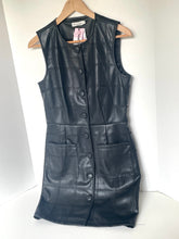 Load image into Gallery viewer, A sleeveless black faux-leather vest dress with a quilted patchwork pattern, button-front closure, and front pockets, hanging on a wooden hanger against a white wall.