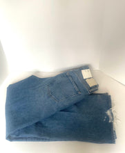 Load image into Gallery viewer, A pair of folded medium-wash blue denim jeans with a raw, frayed hem, displayed on a white corner shelf.