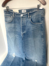 Load image into Gallery viewer, Light blue denim jeans with distressed rips on the lower legs, hanging from a wooden hanger against a white wall.