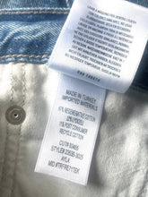 Load image into Gallery viewer, Close-up of a garment's interior labels and stitching, showing white sewn-in care and material labels attached to a light gray fabric body with blue denim-like hem stitching.