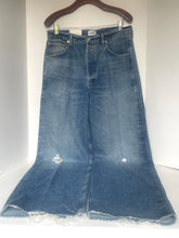 Load image into Gallery viewer, A medium wash blue denim flare skirt with distressed details and raw frayed hems, hanging on a wooden hanger against a white background.
