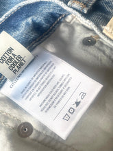 Load image into Gallery viewer, Close-up of a denim garment's interior label area showing a white sewn-in care tag and a side brand tag that reads 'COTTON FOR A COOLER PLANET' and 'CITIZEN OF THE PLANET.'.