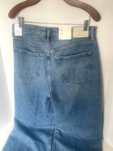 Load image into Gallery viewer, Back view of a pair of medium-wash blue denim jeans hanging on a wooden hanger against a plain white wall.