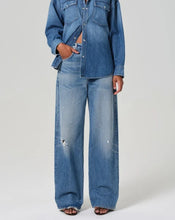 Load image into Gallery viewer, A model wearing a matching blue denim set consisting of a long-sleeve button-up shirt and wide-leg distressed jeans, photographed against a neutral light gray background.
