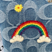 Load image into Gallery viewer, Embroidered rainbow and clouds on denim fabric with a yellow flower.