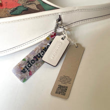 Load image into Gallery viewer, Close-up of a Coach leather ergo handbag with tags and a floral patterned surface.