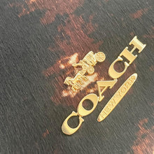 Load image into Gallery viewer, Gold Coach logo charm on a dark wooden surface