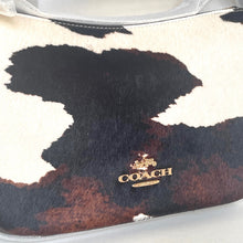Load image into Gallery viewer, Coach handbag with cow print pattern and gold logo on a white background