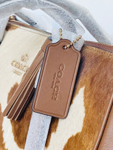 Load image into Gallery viewer, Close-up of a Coach handbag with a brown leather embossed key tag on a chain, next to a cream and brown cowhide-patterned pouch or wallet, with a silver zipper and white protective wrapping visible.