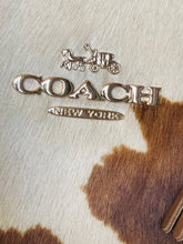 Load image into Gallery viewer, A close-up of a Coach New York logo in polished silver-tone metal, mounted on a brown and white cowhide surface.