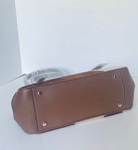 Load image into Gallery viewer, A brown leather handbag shown from the bottom, resting on a light gray surface against a white background, with a clear plastic wrapping still partially visible behind it.
