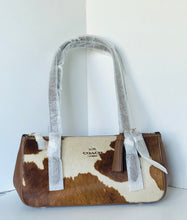 Load image into Gallery viewer, A Coach handbag with a brown and cream cowhide pattern, featuring silver-gray handles still wrapped in protective plastic, set against a plain white corner wall.