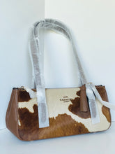 Load image into Gallery viewer, A Coach shoulder bag with a brown and cream cowhide pattern, brown leather trim, and protective clear plastic wrapping on the handles, set against a plain white background.