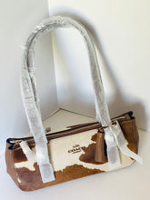 Load image into Gallery viewer, A Coach shoulder bag with a brown and white cow print pattern, featuring a faux-leather strap wrapped in protective white paper and a matching brown leather tassel charm.