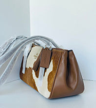 Load image into Gallery viewer, A Coach handbag with a brown leather and brown-and-white cowhide pattern, featuring a brown leather tassel tag with the Coach logo, resting on a light gray surface against a white background.