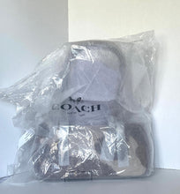 Load image into Gallery viewer, A Coach handbag wrapped in a clear plastic protective bag, with the Coach logo visible on the front label, set against a plain white background.
