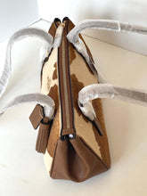 Load image into Gallery viewer, A brown and beige cowhide handbag with a brown leather tassel and a gold-tone zipper, shown on a white surface with protective white foam wrapping around its handles.