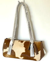 Load image into Gallery viewer, A Coach handbag with a brown and cream cowhide pattern, featuring a silver-tone shoulder strap wrapped in protective plastic and a brown leather tassel.
