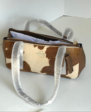 Load image into Gallery viewer, A Coach shoulder bag with a brown and cream cowhide pattern, featuring a brown leather trim, gold-tone zipper, and a tassel charm. The handles are still wrapped in protective white plastic packaging, and the bag is photographed on a plain white surface.