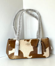 Load image into Gallery viewer, A Coach shoulder bag with a brown and cream cowhide pattern, featuring protective plastic wrapping on the handles and a visible Coach logo on the front.