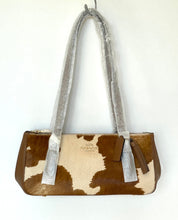 Load image into Gallery viewer, A Coach handbag with a brown and cream cowhide pattern, featuring silver-gray velvet handles wrapped in protective plastic and a small brown leather brand tag.