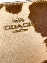 Load image into Gallery viewer, Close-up of a Coach New York handbag featuring a brown and cream cowhide pattern with a polished gold-tone metal logo on the front.