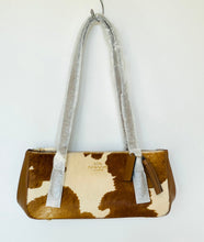 Load image into Gallery viewer, A Coach handbag with a brown and cream cowhide pattern, brown leather trim, and silver-gray straps, shown hanging against a plain white wall.