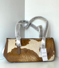 Load image into Gallery viewer, A Coach handbag with a brown and cream cowhide pattern, featuring protective gray plastic wrapping on the handles and a white product tag hanging from the zipper.