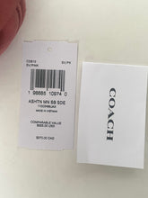 Load image into Gallery viewer, A Coach garment tag and a white Coach-branded card on a light surface, with part of a finger visible at the top left.