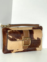 Load image into Gallery viewer, A wristlet clutch bag featuring a brown and beige cowhide pattern with tan faux-leather trim, a front flap pocket with a gold-tone buckle, and a top handle, shown against a white background.