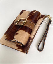 Load image into Gallery viewer, A small wristlet clutch with a brown and beige cowhide pattern, a brown strap with a gold buckle, and a matching wrist strap, shown on a plain light background.