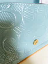 Load image into Gallery viewer, Close-up of a light blue embossed leather clutch or wallet featuring the signature Coach 'C' monogram pattern and a gold-tone snap button closure.